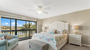 Un dormitorio con una cama grande, ventilador de techo, mesita de noche, lámpara, banco y vista a palmeras a través de amplias ventanas.
