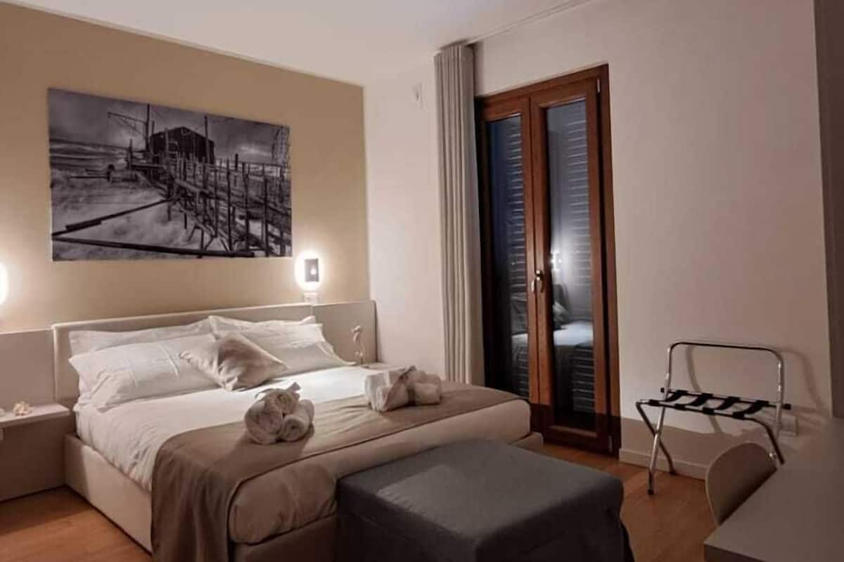 A hotel room with a bed, a small table, a chair, a painting on the wall, and a door with a view.
