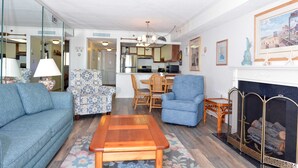 A living room with a sofa, armchair, and coffee table, a dining area with chairs, and a fireplace. - This two-bedroom, two bathroom condo has a beautiful view of the ocean, and outdoor pool. (Ocean City)