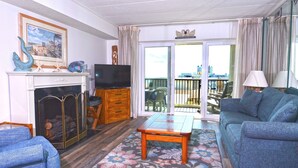 A living room with a fireplace, a sofa, a television, and a balcony with a city view. - This two-bedroom, two bathroom condo has a beautiful view of the ocean, and outdoor pool. (Ocean City)