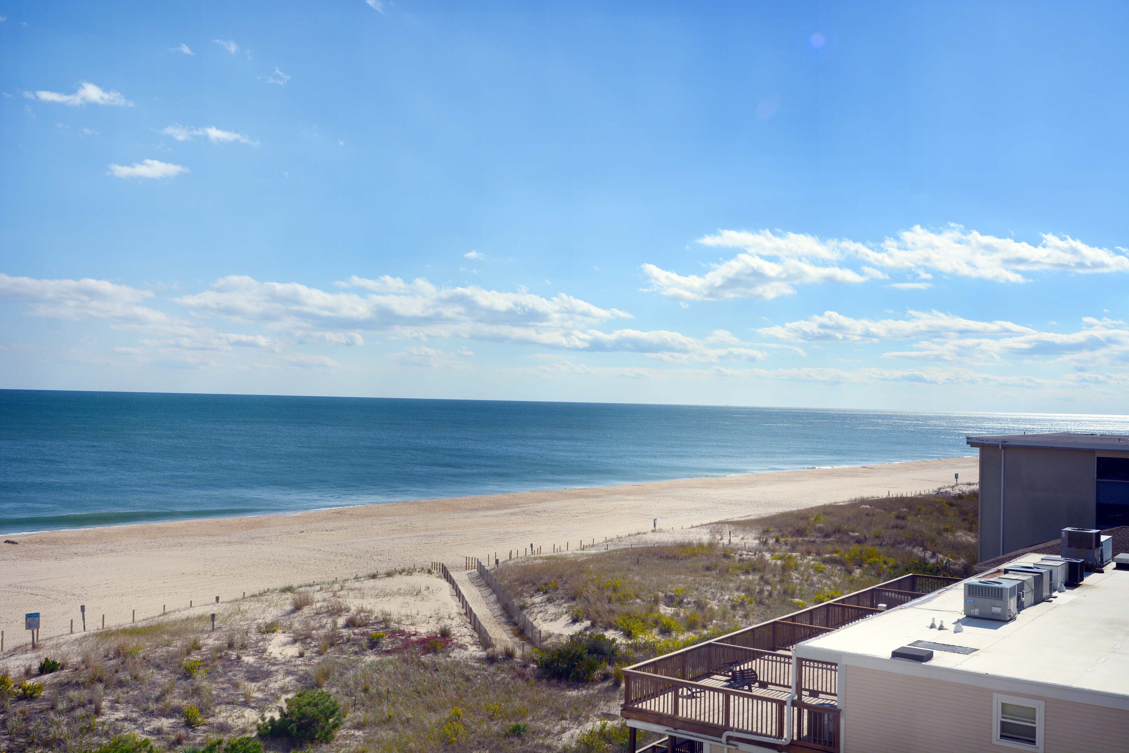 A beach view with a sandy shore, a wooden fence, and a building with air conditioning units.