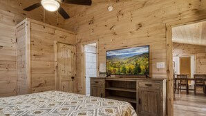 A wooden cabin bedroom with a bed, a television on a wooden stand, and a view of a forest through a window.