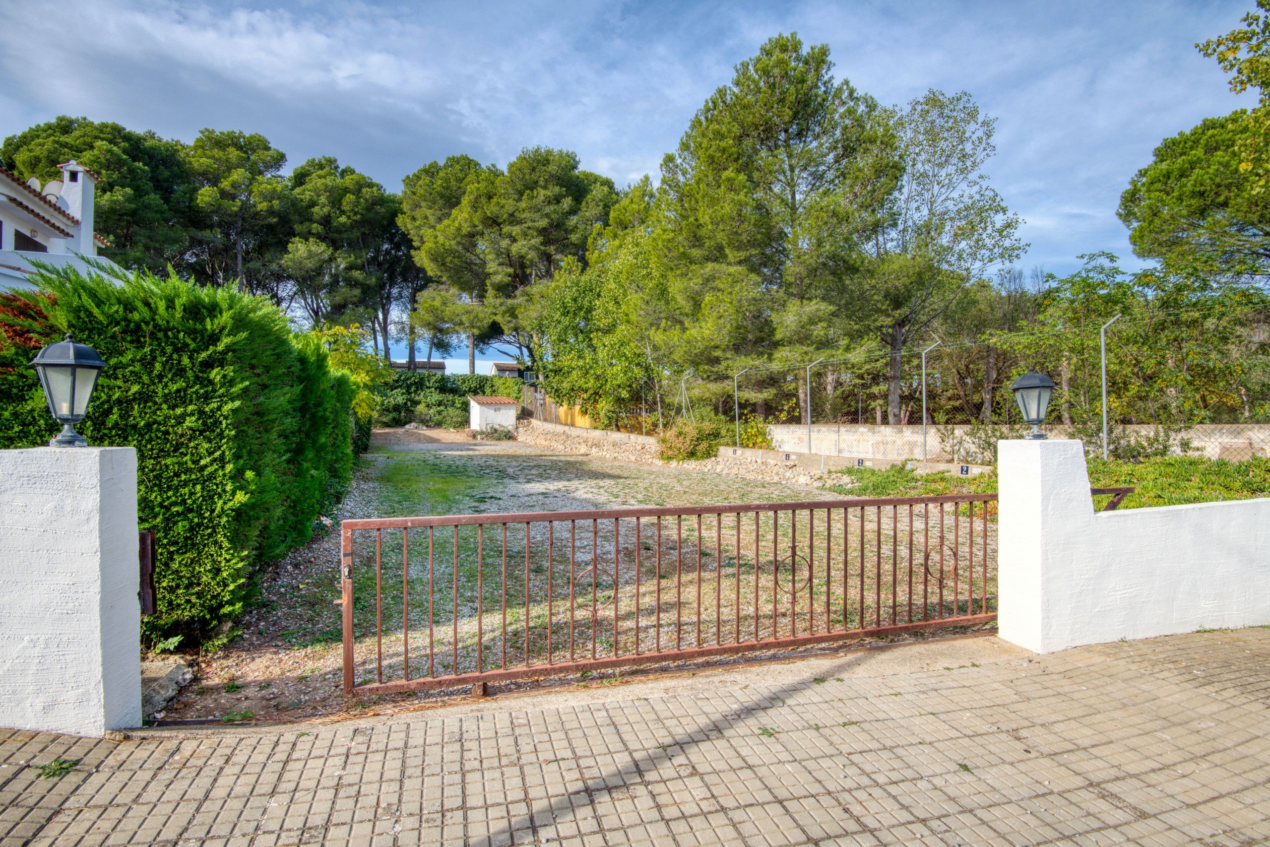 A paved pathway leading to a gated entrance with a metal fence, surrounded by lush greenery and trees.