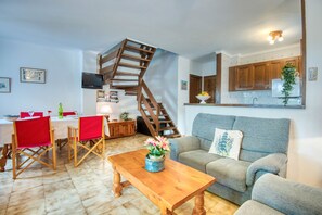 Sala de estar com sofá cinza, mesa de centro de madeira e área de jantar com cadeiras vermelhas. Uma escada espiral de madeira leva a um andar superior.