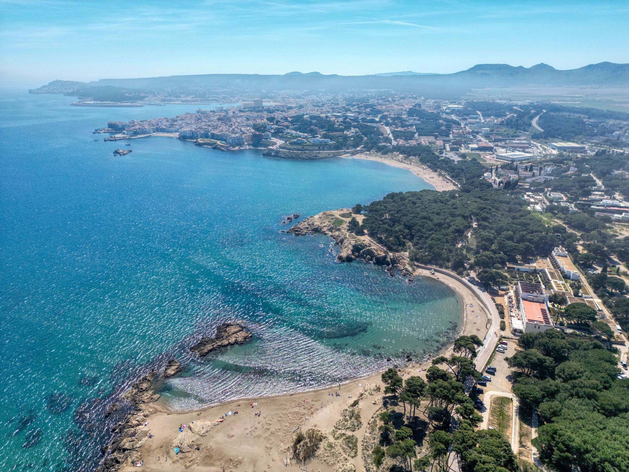 Une vue aérienne d’une zone côtière, avec une plage, des eaux d’un bleu limpide et un mélange de paysages urbains et naturels.