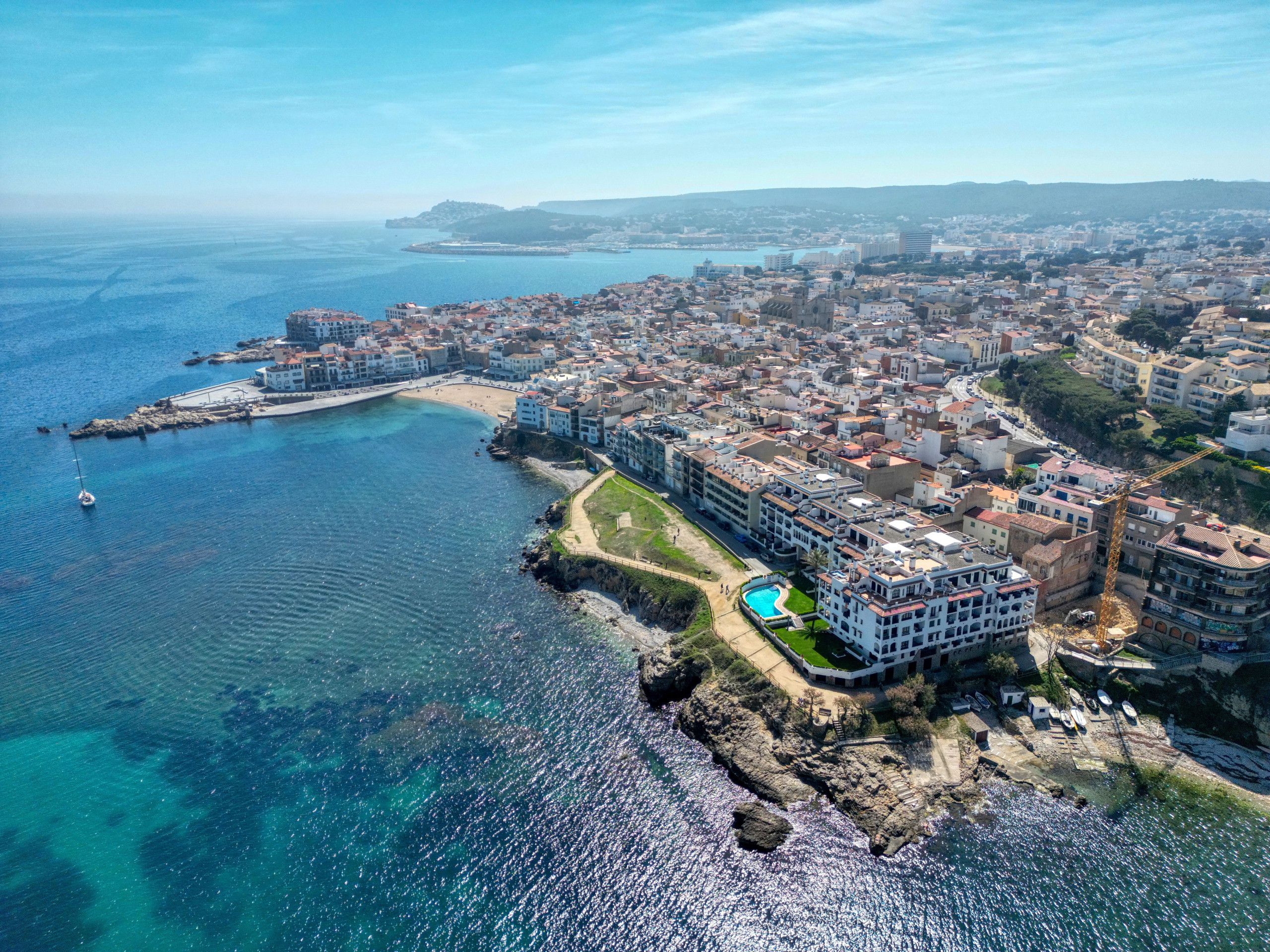 An aerial view of a coastal town with a marina, buildings, and a clear blue sea.