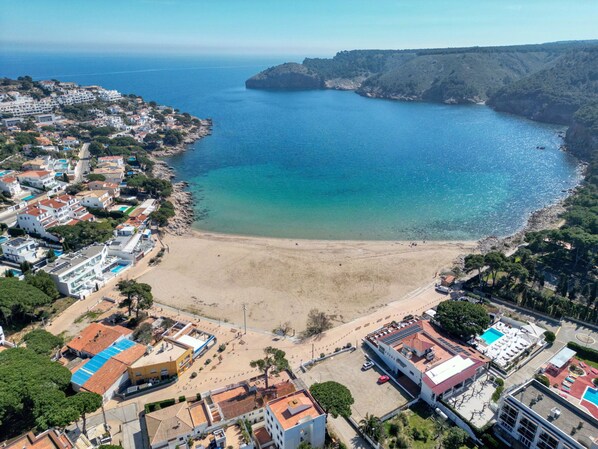 An aerial view of a coastal town with a sandy beach, residential buildings, and a clear view of the sea.