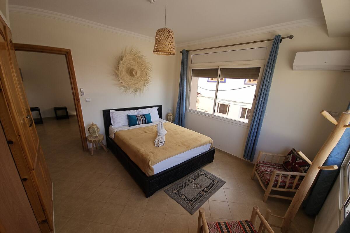 A bedroom with a bed, a window with curtains, a hanging light fixture, a wooden chair, and a small table.