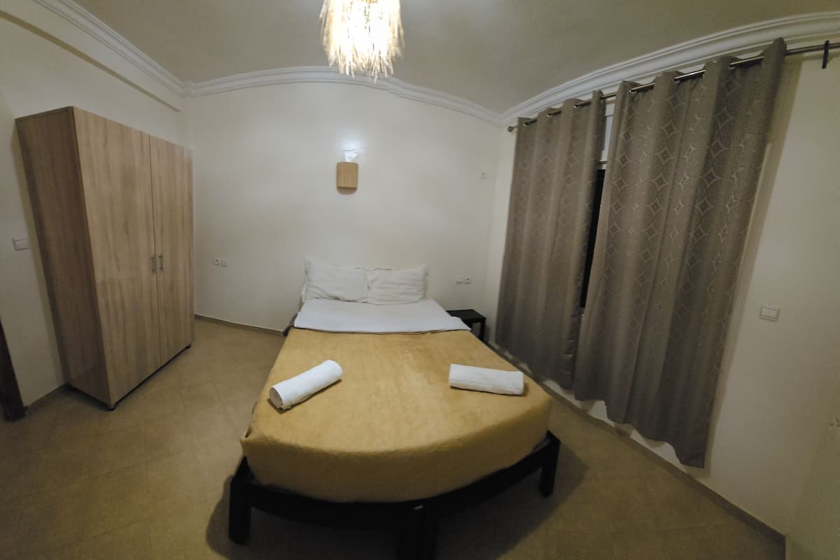A single bed with a beige mattress and white linens, a wooden wardrobe, and a small bedside table.