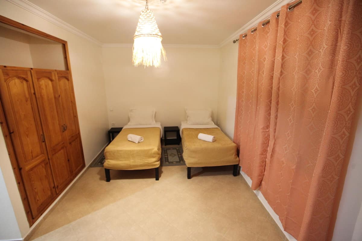 A room with two single beds, a wooden wardrobe, a small table, and a patterned curtain.