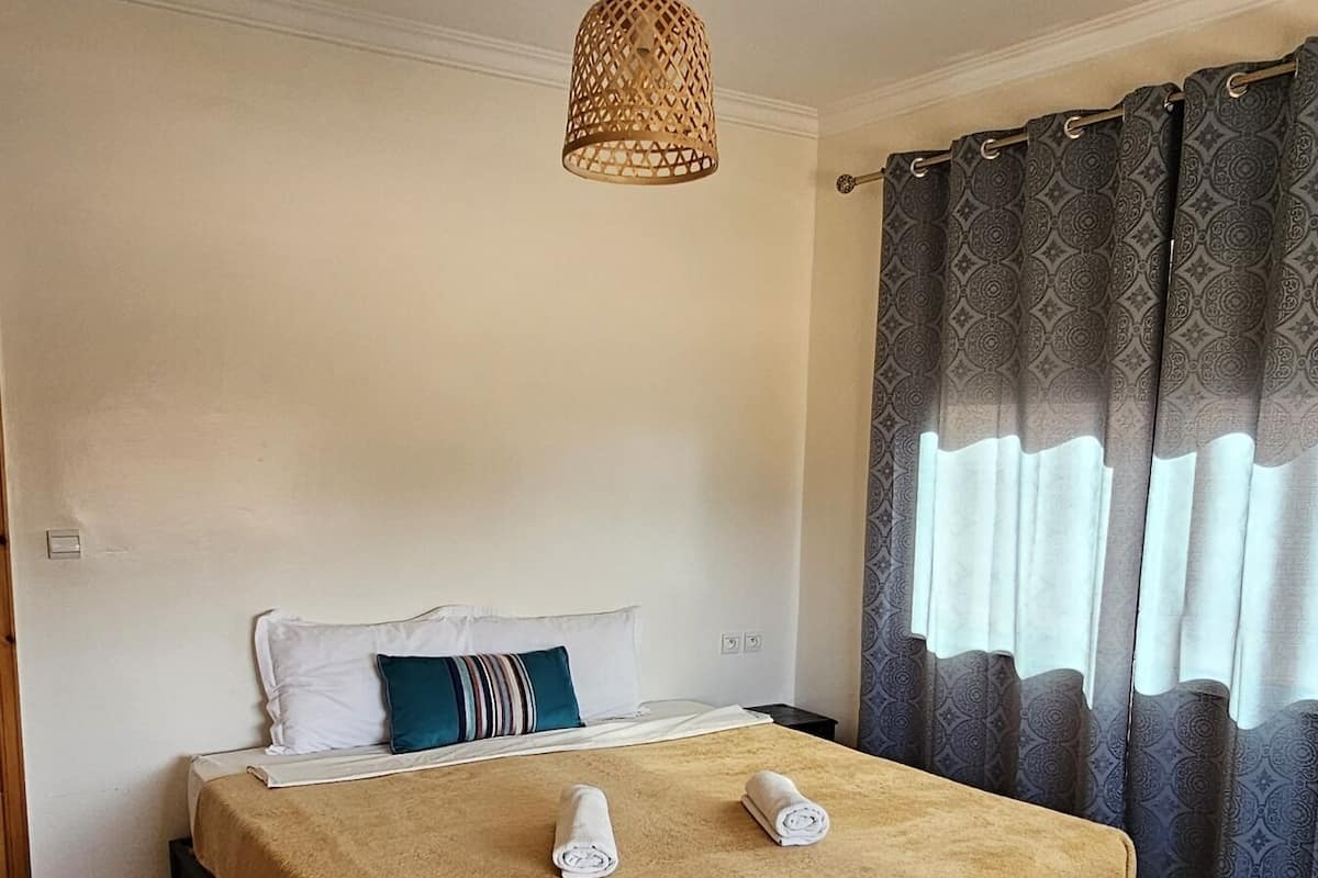 A neatly made bed with a brown blanket, a hanging rattan light fixture, and a window with patterned curtains.