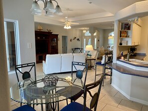 Un vaste salon avec un canapé blanc, une table à manger en verre entourée de chaises, un coin cuisine doté d’un évier et d’un plan de travail, et un ventilateur de plafond.