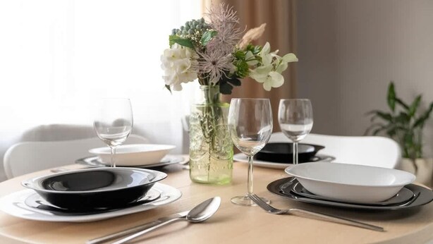 Una mesa de comedor preparada con platos, cubiertos y vasos, acompañada de un jarrón con flores.