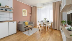 Una sala de estar moderna con área de cocina, piso de madera, un sofá gris, una mesa de comedor con sillas y un gran ventanal con cortinas.