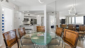 A modern dining area with a glass table surrounded by wooden chairs, a kitchen in the background, and a large window providing natural light. - ⭐Newly Remodeled Beach FrontCondo-Great Views from Balcony-Windward Pointe 603⭐ (Orange Beach)