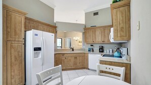 A modern kitchen with wooden cabinets, a white refrigerator, a sink, and a dining area with a white table and chair. - Valencia in Pelican Bay (Naples)