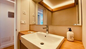 Une salle de bain avec un lavabo blanc, un miroir, un plan de toilette en bois et un flacon de lotion.