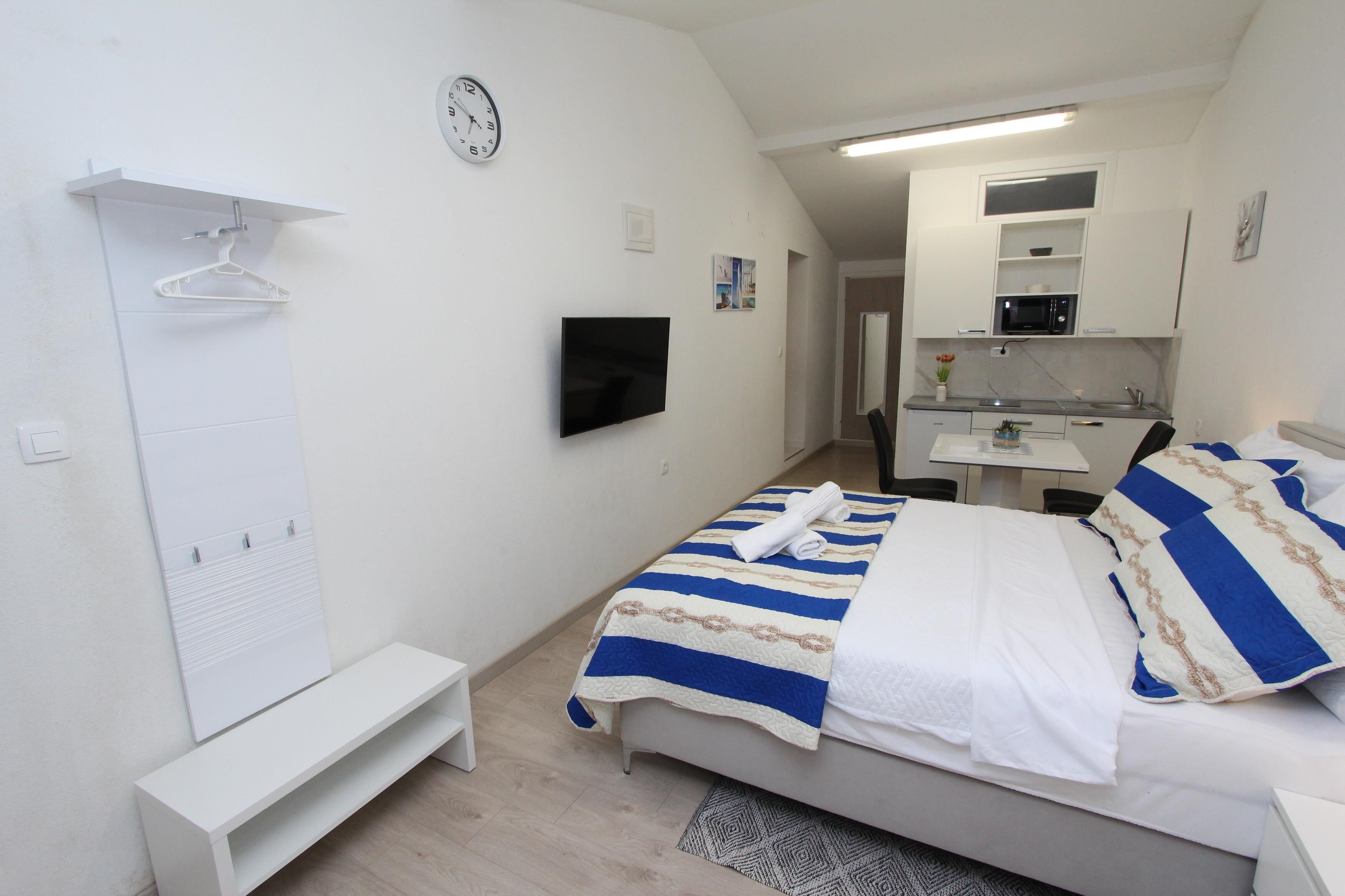 A bedroom with a bed, a TV, a desk, and a kitchenette.