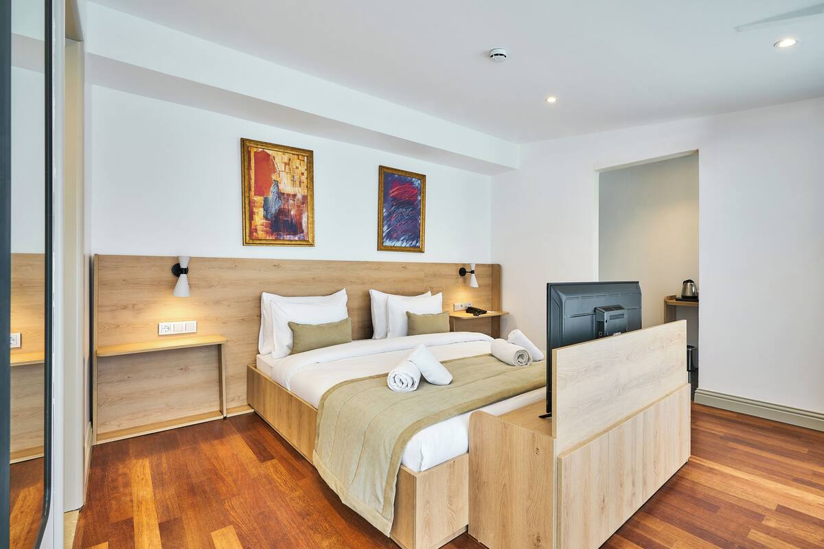 A modern hotel room with a wooden headboard, a bed with white linens, two wall-mounted paintings, a bedside table with a lamp, and a TV on a wooden stand.