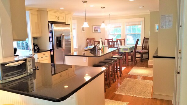 A modern kitchen with a central island, stainless steel appliances, and a dining area with wooden chairs and a table. - Short Distance to Colonial Acres Beach (2342) (West Yarmouth)