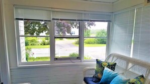 A room with a wicker sofa and a table, a large window with blinds, and a view of a green landscape. - Short Distance to Colonial Acres Beach (2342) (West Yarmouth)