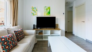 Una sala de estar moderna con un sofá, una mesita de centro y un televisor sobre un mueble de madera. Hay dos obras de arte en la pared y una puerta al fondo.