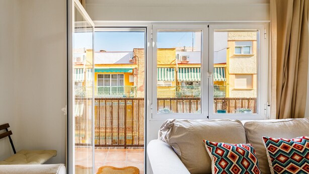 Un salón acogedor con un sofá, cojines coloridos y un gran ventanal corredero que da a un balcón con vistas a edificios de colores.