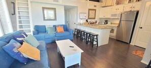 A living room with a blue sofa, a white coffee table, and a kitchen area with a refrigerator and bar stools. - Oyster Cove (Port Isabel)