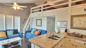 A compact living space with a kitchenette, a blue sofa, a ceiling fan, and a staircase to an upper level. - Bay Sunset Views - Waterfront Pool - Boat Slip (Port Isabel)