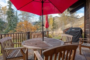 Une terrasse en bois avec une table ronde et des chaises, un parasol rouge et un barbecue.