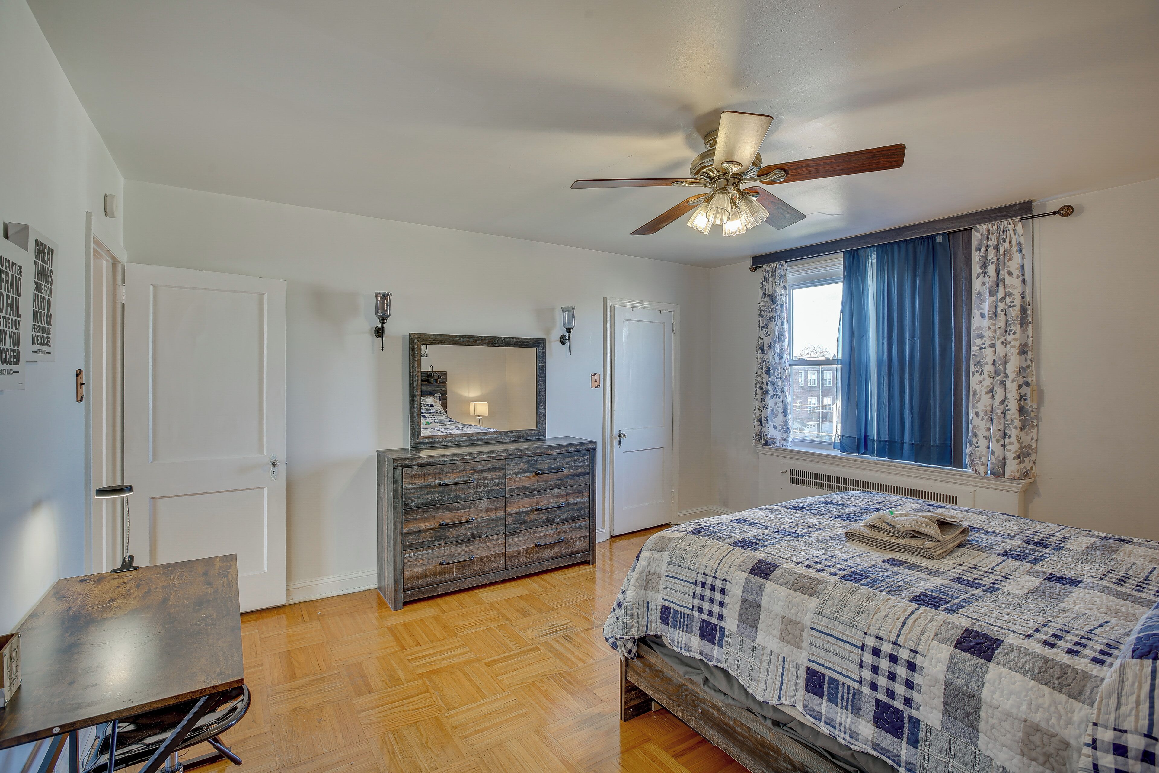 A bedroom with a wooden floor, a bed with a quilt, a dresser, a ceiling fan, and a window with curtains.