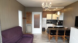 Een ruimtelijk ingerichte leefruimte met een paarse bank, een eettafel met stoelen, een keuken met apparatuur, en een tv op een standaard.