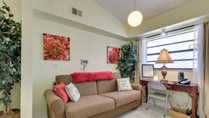 A living room with a brown sofa, red and floral cushions, a desk with a lamp, and a computer. There are two large floral paintings on the wall and a potted plant to the left. - Orlando Home: 6 Mi to Universal! (Orlando)
