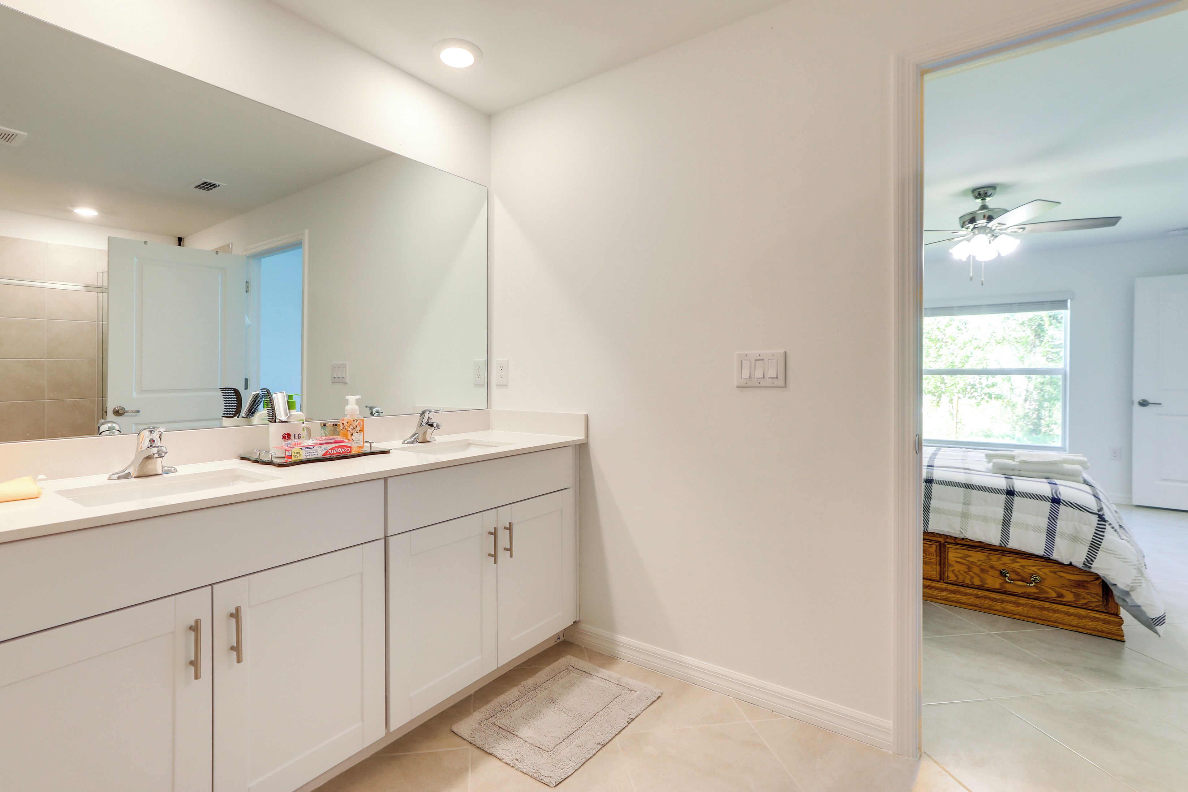 A modern bathroom with a large mirror, two sinks, and a wooden bed in the adjacent room.