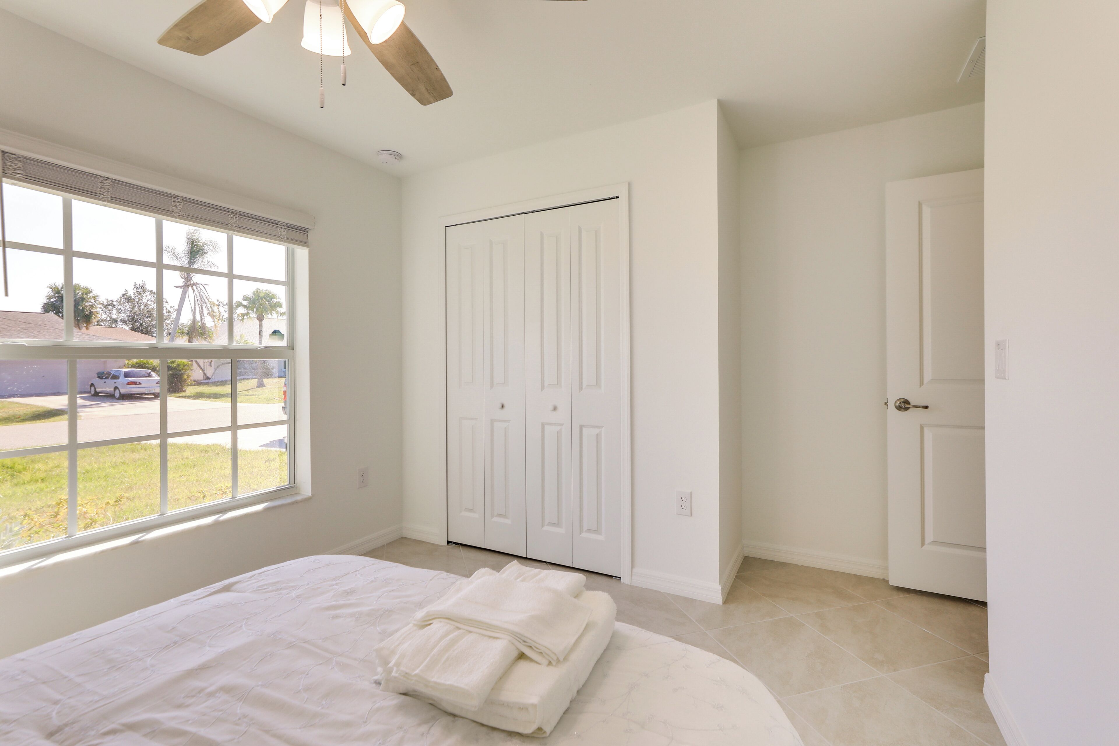 A bedroom with a large window, a bed with neatly folded white linens, and a white wardrobe.