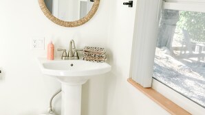 Un baño con lavamanos pedestal blanco, un espejo redondo con marco de mimbre, un perchero y una ventana con vista a árboles.