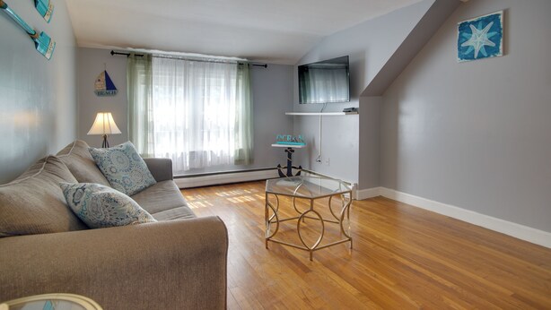 Un salón con un sofá, una mesa de centro de vidrio, una lámpara, un televisor y una ventana con cortinas.