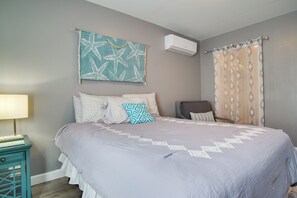 A bedroom with a bed, pillows, a chair, a wall-mounted air conditioner, and a curtain. - Central Cape Cod Escape in South Yarmouth! (South Yarmouth)