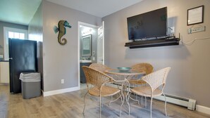 Un coin repas avec des chaises en osier et une table, un téléviseur fixé au mur et un réfrigérateur à l’arrière-plan.