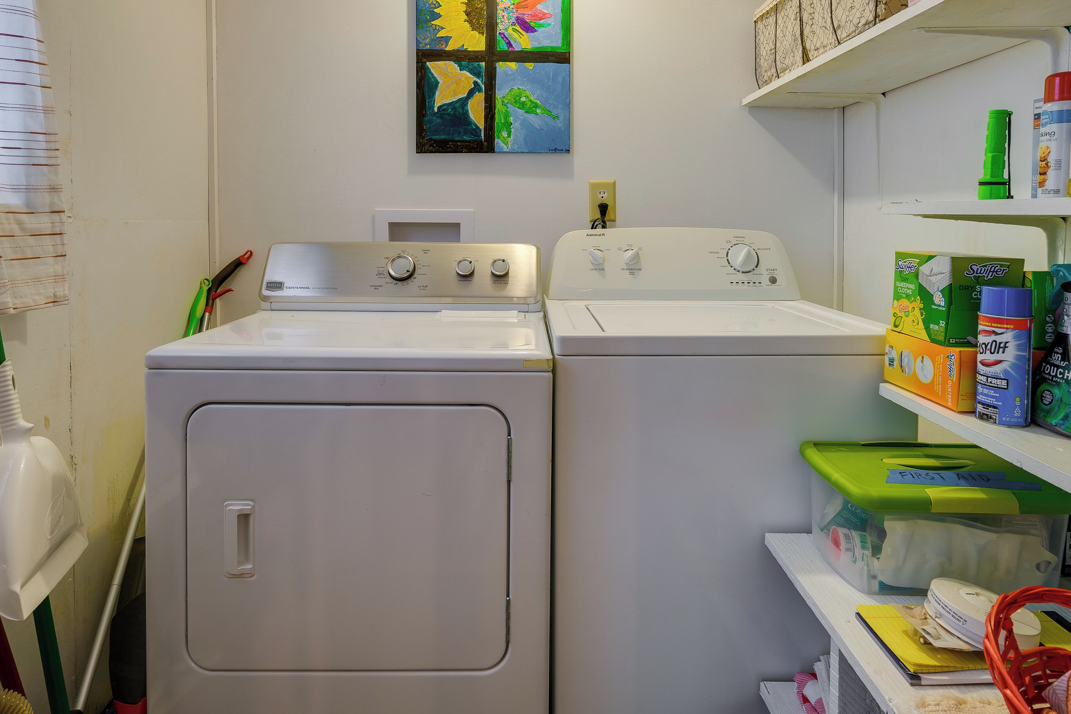 A laundry area with two white washing machines, a shelf with cleaning supplies, and a colorful artwork on the wall.