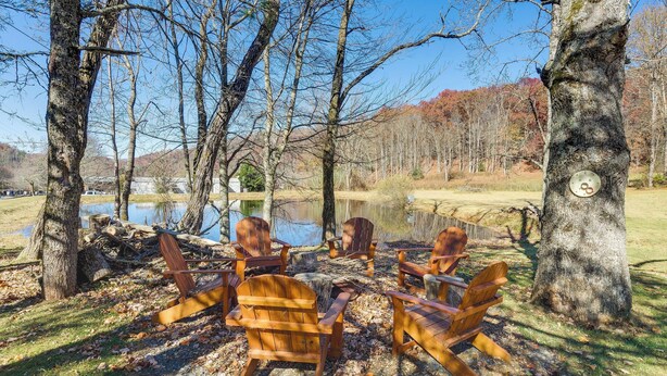A lakeside setting with wooden Adirondack chairs arranged around a table, surrounded by trees and a clear sky. - Pet-friendly Topton Home w/ Patio, Deck & Views! (Topton)