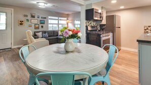 A dining area with a round table and four chairs, a vase with flowers, a sofa, and a kitchen in the background.