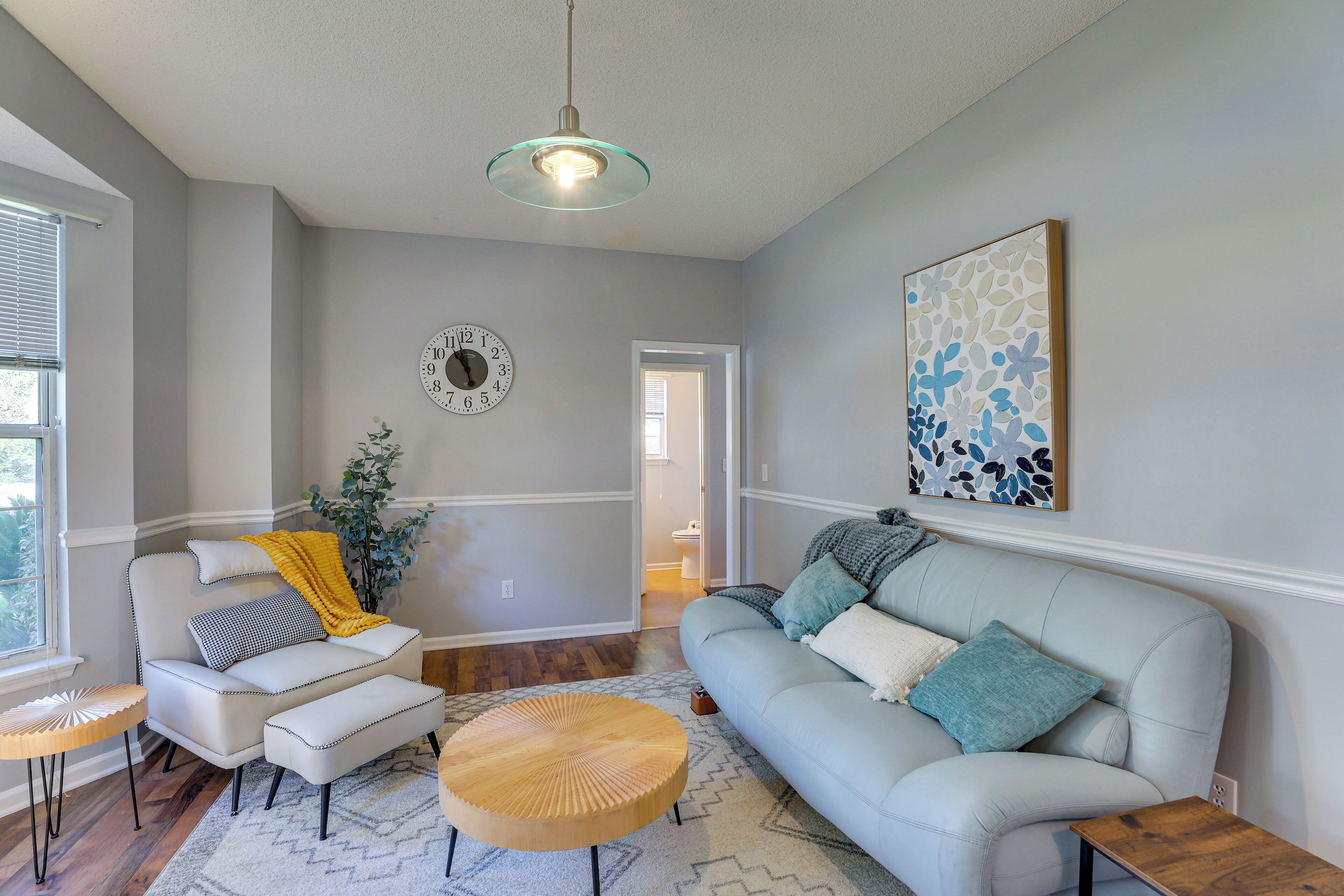 Un salon avec un canapé bleu clair, une table basse ronde en bois, un fauteuil blanc et une petite table d’appoint. On peut voir une grande horloge murale et un tableau d’art abstrait encadré.