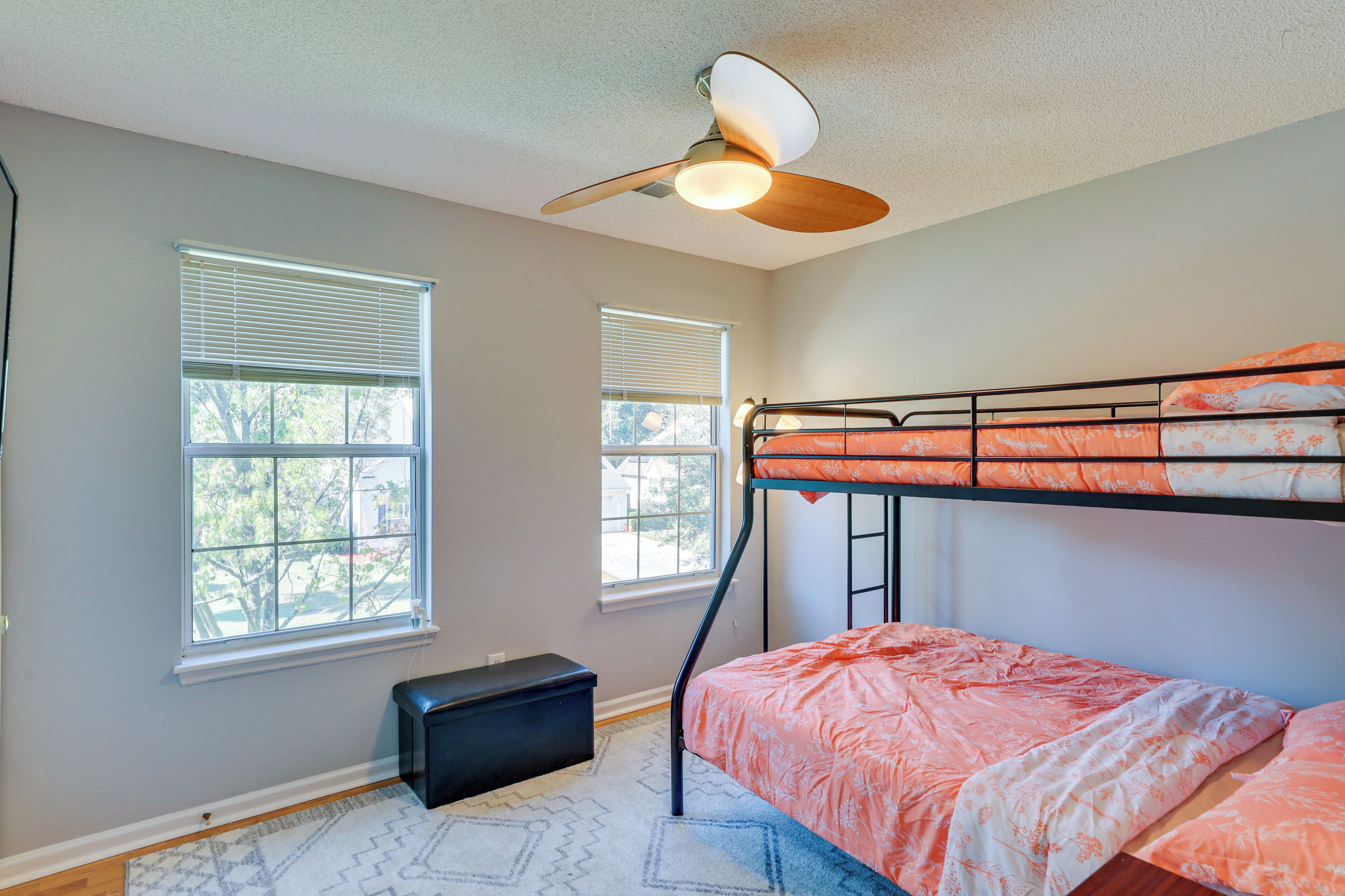 A bedroom with a bunk bed, two windows with blinds, a ceiling fan, and a small black bench.