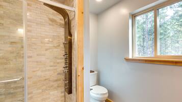 Un baño con cabina de ducha de vidrio, inodoro y ventana con vista a árboles.