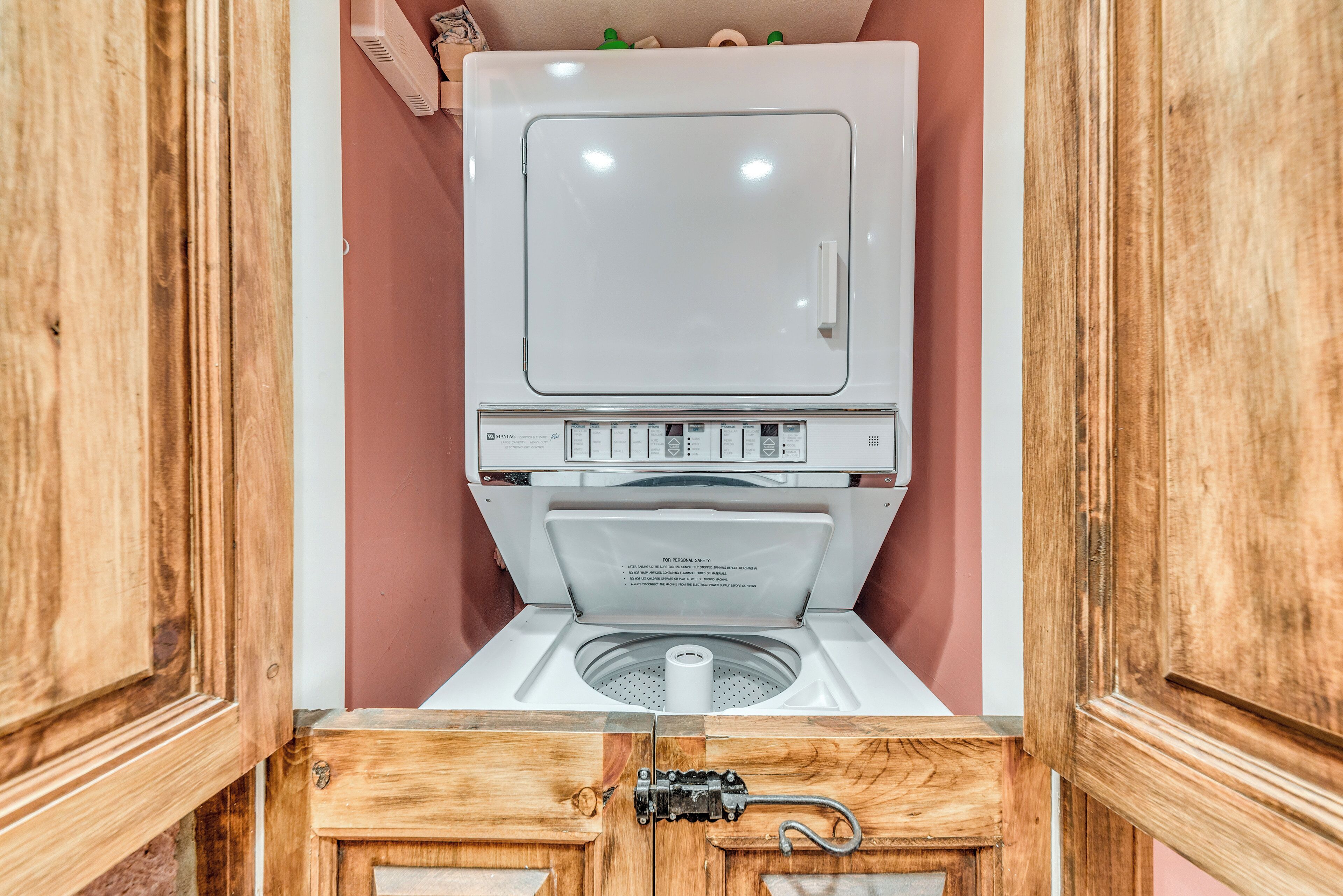 Une machine à laver compacte, blanche, à chargement frontal, intégrée dans un meuble en bois, avec un mur rose en arrière-plan.