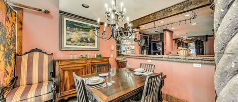 A dining area with a wooden table set for four, surrounded by upholstered chairs. A chandelier hangs above the table, and a painting is visible on the wall.