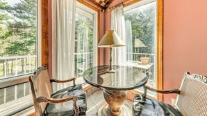 Una habitación con una mesa redonda de cristal, dos sillas con cojines de cuero, una lámpara de latón y amplias ventanas con cortinas.