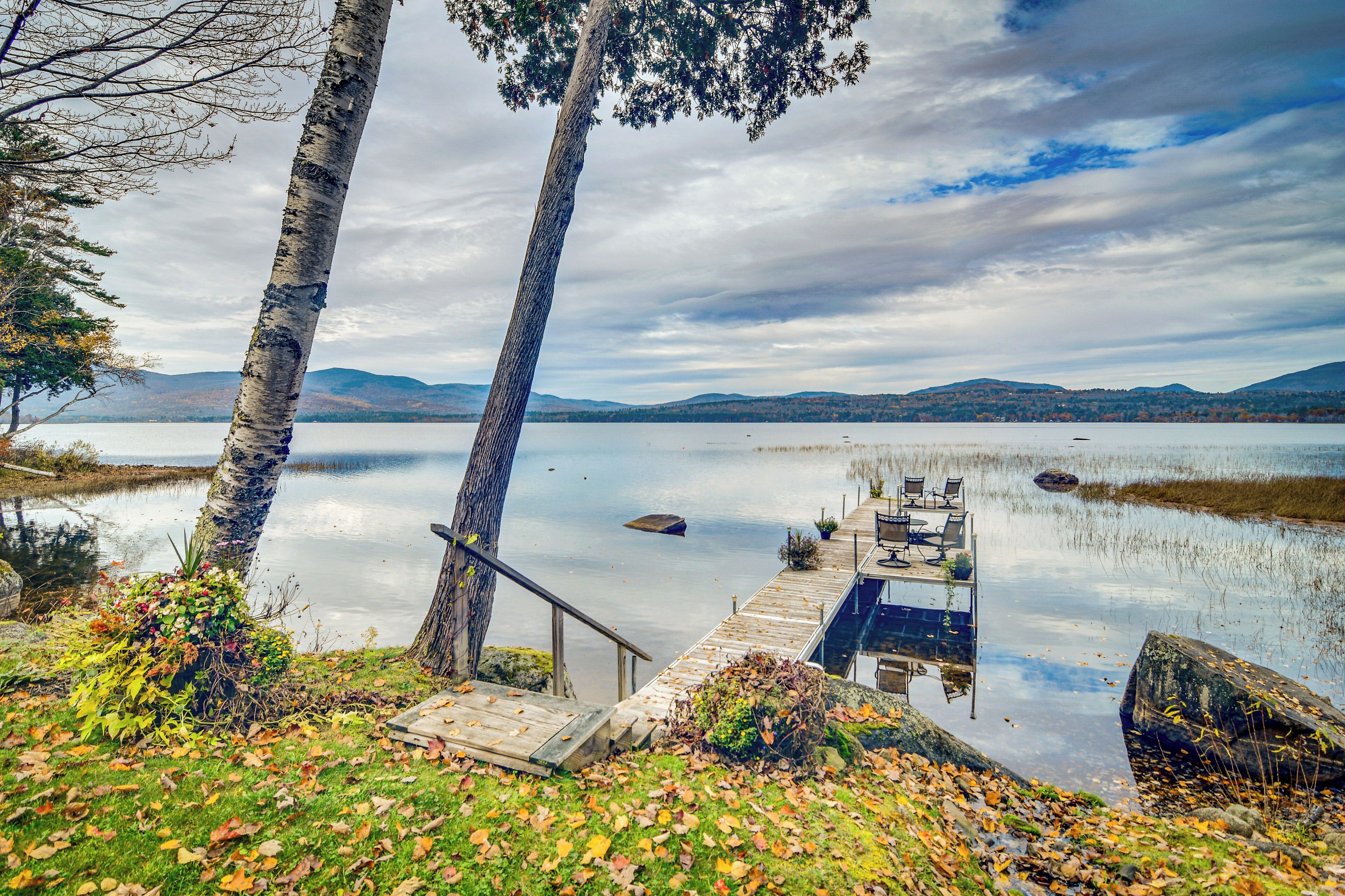 A lakeside dock with a wooden walkway leading to a dock chair, surrounded by trees and a calm lake with distant mountains.