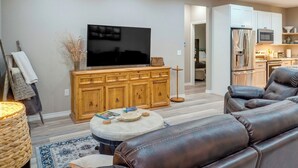 A modern living room with a flat-screen TV, leather sofas, a wooden cabinet, and a kitchen area in the background.